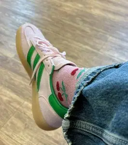 A pair of pink suede trainers with the signature adidas stripes i green worn with pink glittery socks with red cherries on and cropped jeans